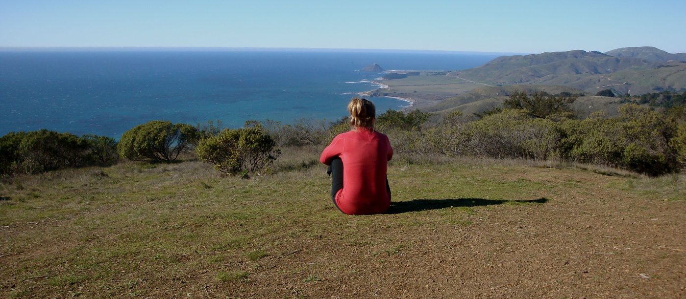 A person sitting on a grass headland above the coast, looking out to sea